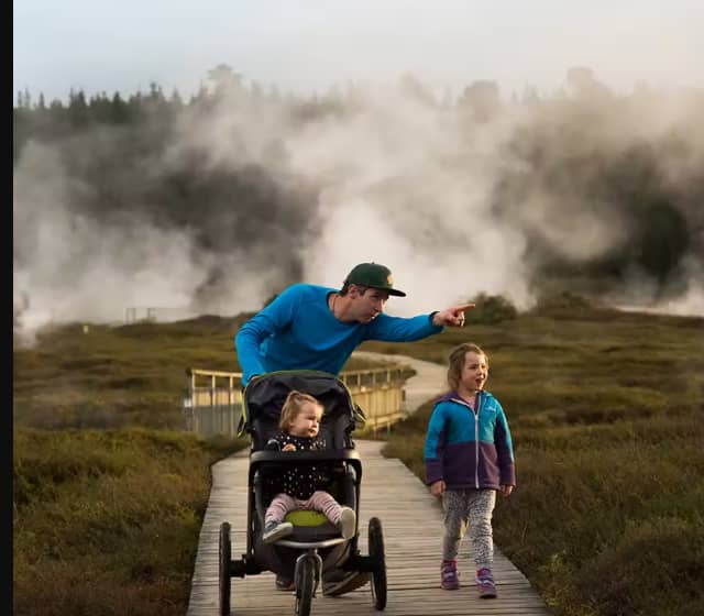 Man with two children walking on a boardwalk with geothermal steam in the background.
