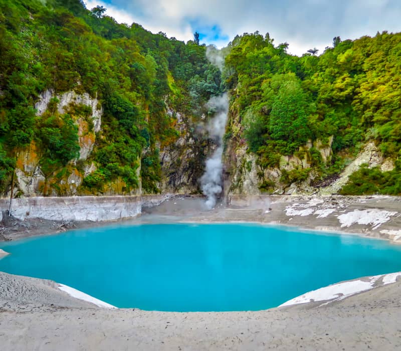 A bright turquoise geothermal lake, Inferno Crater, sits at the base of a steep, forest-covered cliff with a plume of white steam rising from the water's edge at Waimangu Volcanic Valley.
