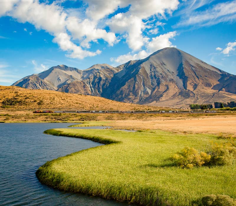 TranzAlpine train passing near Lake Sarah with mountains and blue summer sky