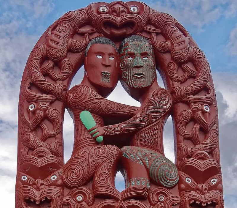 Red Maori wood carving of two figures intertwined with intricate patterns under a cloudy sky