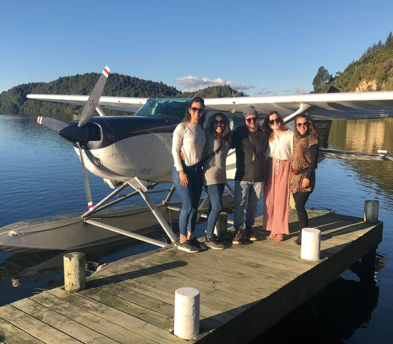 Five people standing on a dock next to a seaplane on a sunny lake with forested hills.