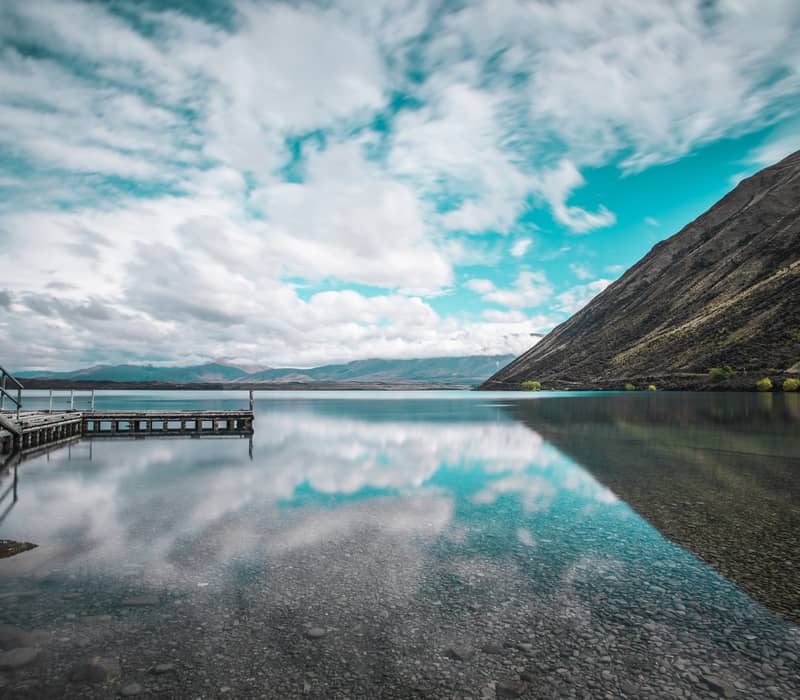 Clear lake with mountain slopes, wooden pier, and clouds reflected in the water at Lake Ohau.