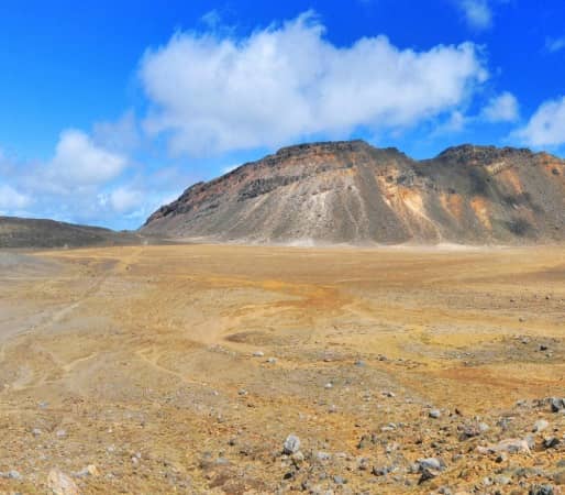 A wide panoramic view of the Tongariro Alpine Crossing showing hikers walking through a barren, rocky volcanic crater with the towering cone of Mount Ngauruhoe in the background under a blue sky.