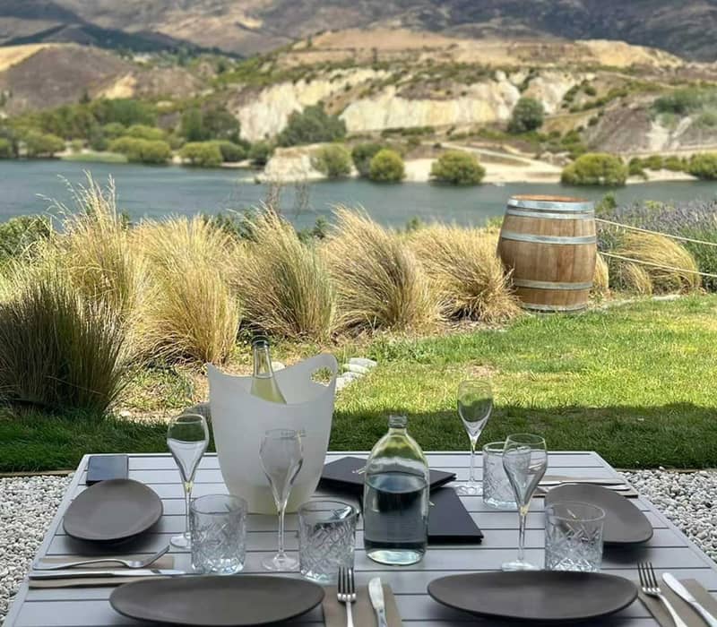 Outdoor table set with wine glasses and plates by a lake with hills in the background.
