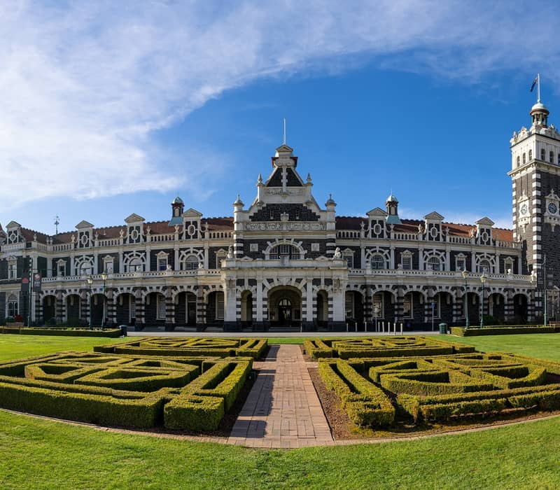Historic Dunedin Railway Station with clock tower and neatly trimmed hedge garden under clear blue sky.