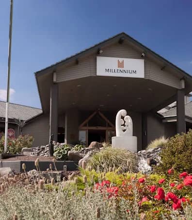 Entrance of Millennium Hotel Rotorua with sculpture and colorful garden under a partly cloudy sky.