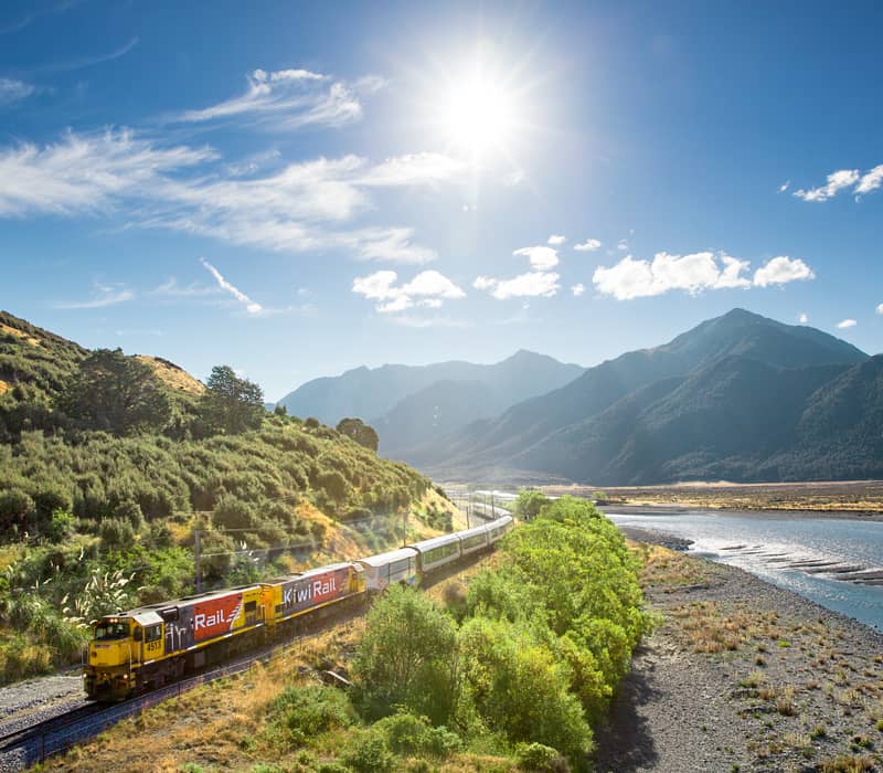 Scenic train crossing a bridge over the Waimakariri River with mountains in the background on a clear day.