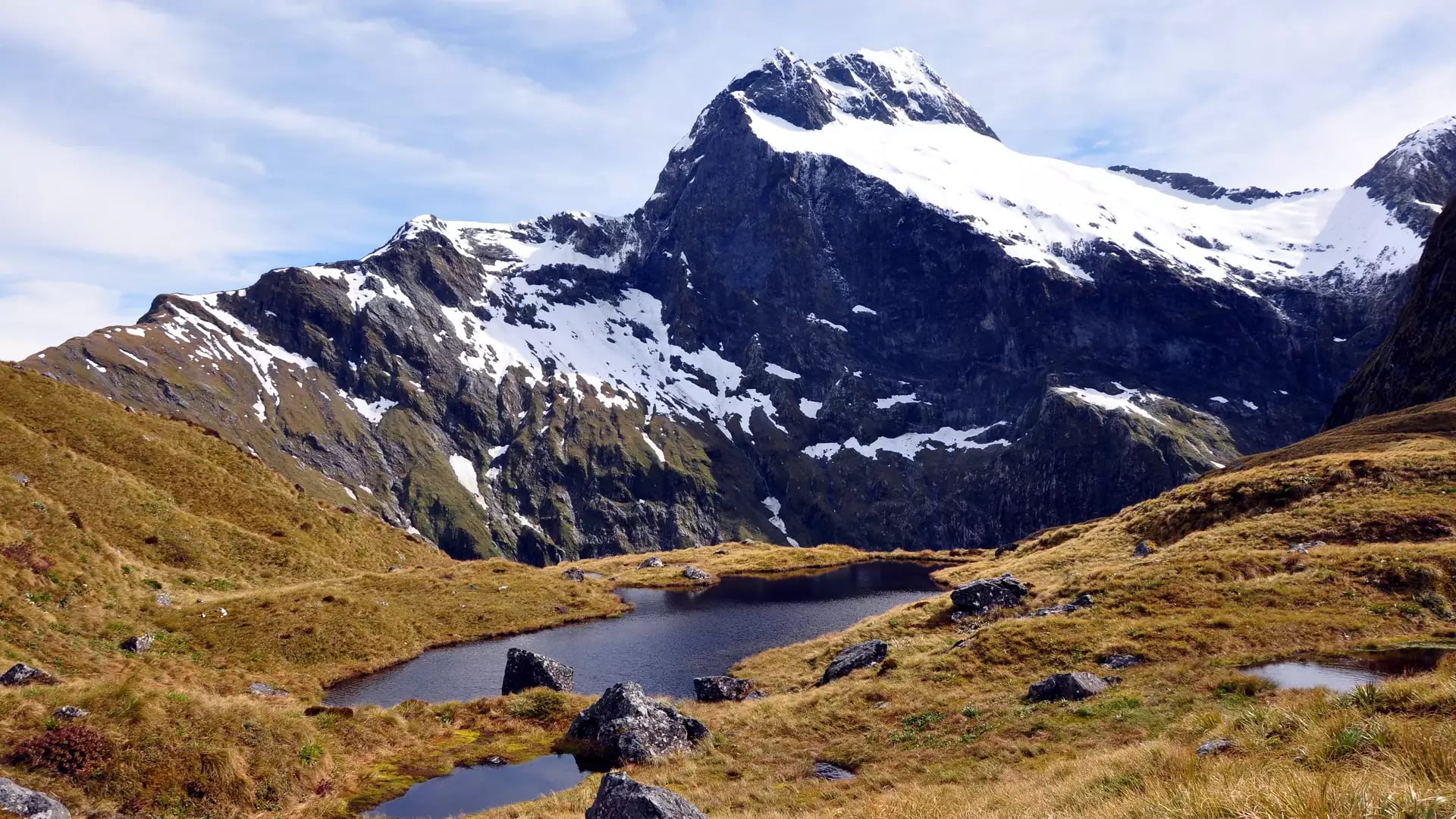 Snow-capped mountain behind a small alpine lake with grassy and rocky terrain.