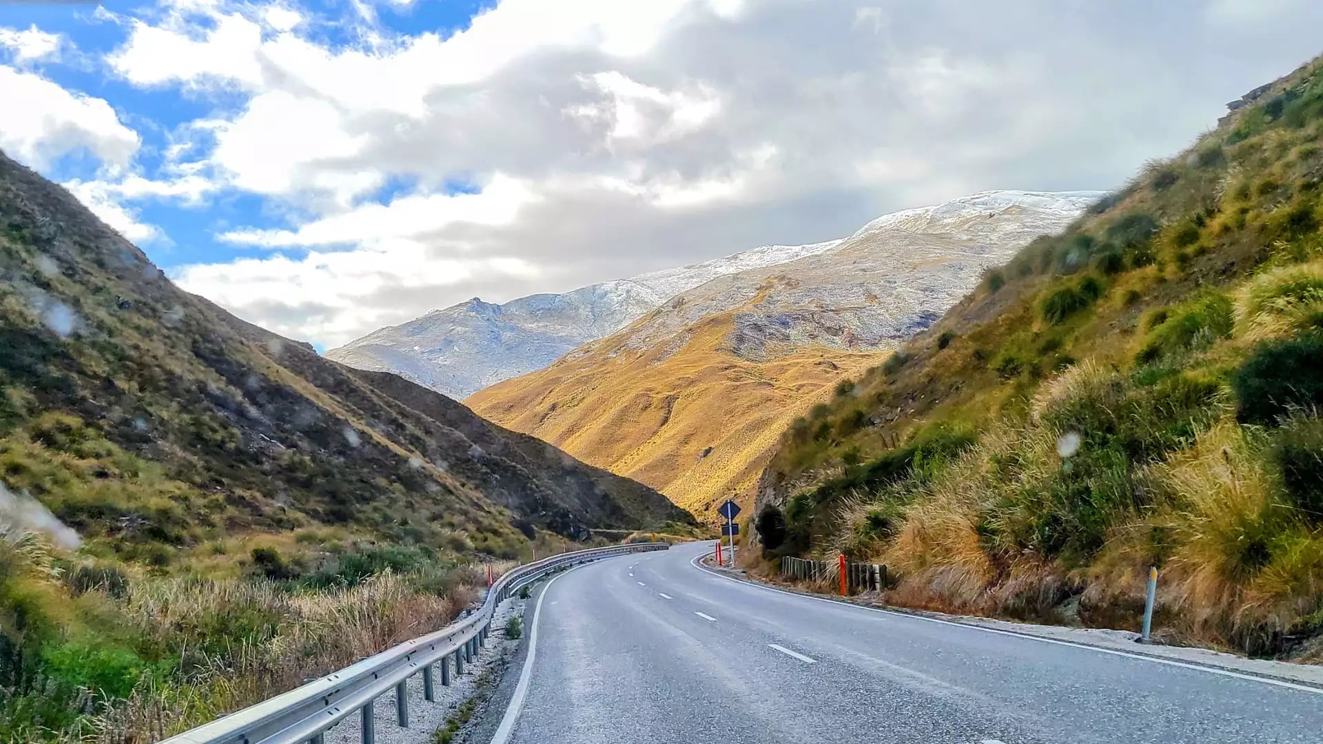 Winding mountain road through grassy hills and snow-capped peaks under a cloudy blue sky.