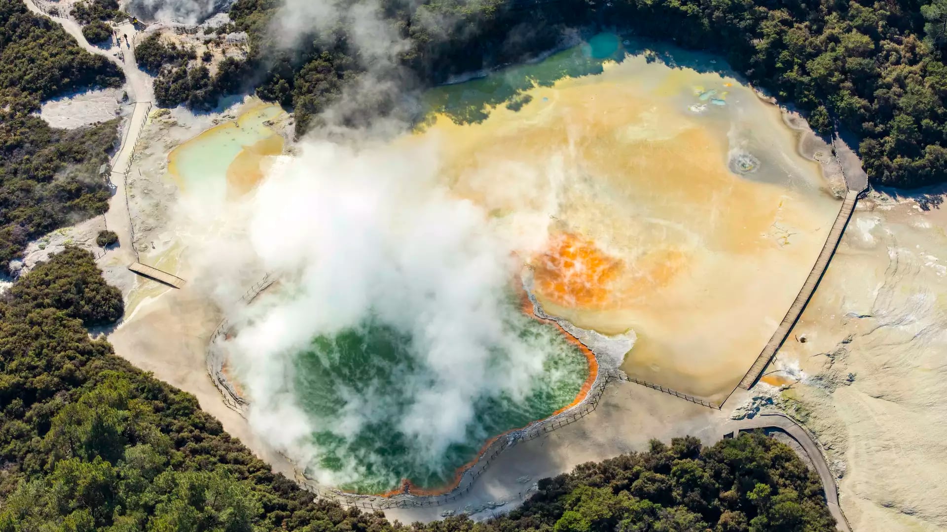 Aerial shot of a colorful geothermal hot spring emitting steam with forest and boardwalks nearby