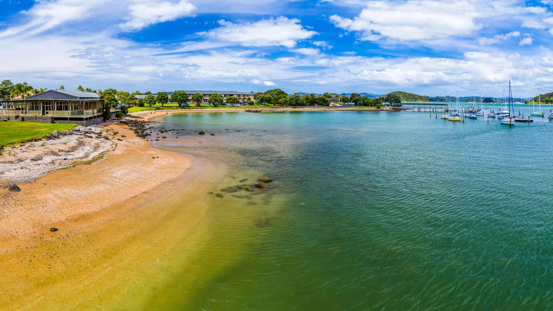 A panoramic view of Te Ti Bay in Paihia, showing a golden sand beach, clear green-blue water with moored boats, and beachfront buildings under a bright blue sky.
