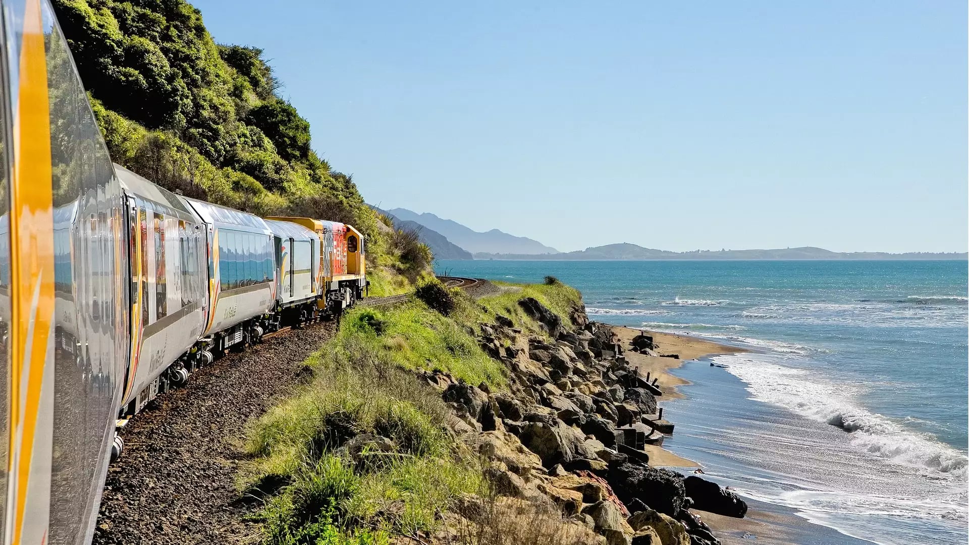 Train traveling along a coastal railway with the ocean and beach nearby under a clear sky.