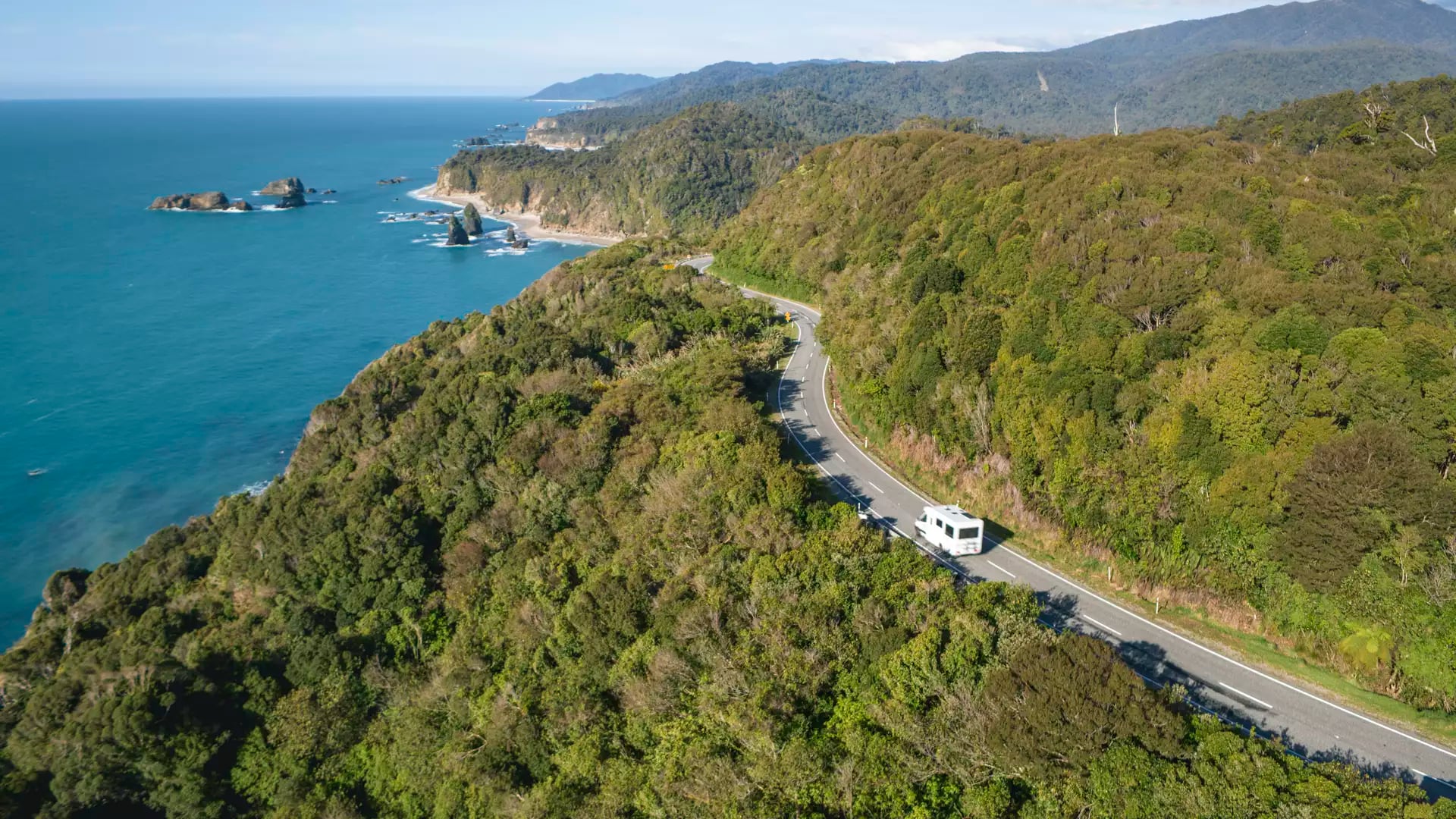 An aerial view of a white campervan driving along a winding coastal road bordered by dense green rainforest and the turquoise Tasman Sea.