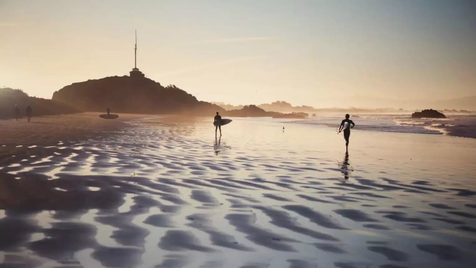 Surfers walking on a beach at sunset carrying surfboards with rippled sand and waves in the background.