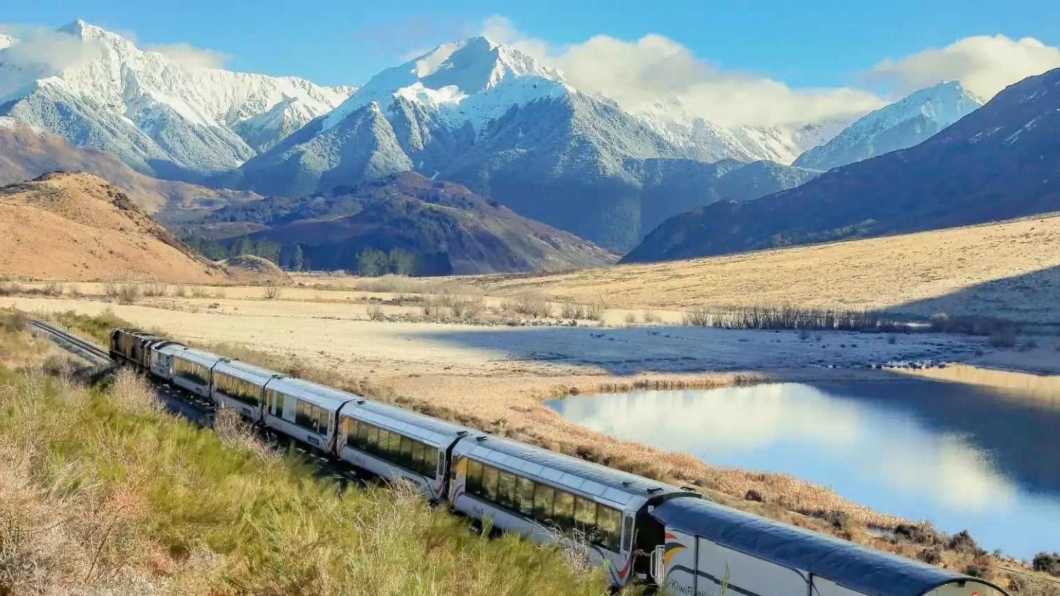 Train traveling through a mountain valley with snow-covered peaks and a calm lake.