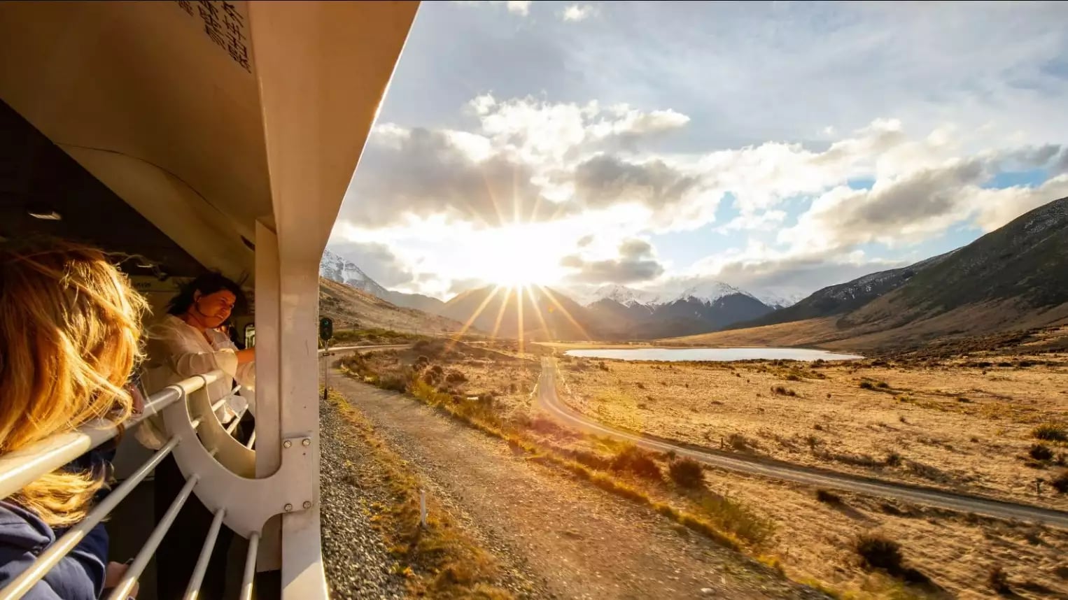 People on a train looking at a sunset over mountains and a lake in a wide valley.