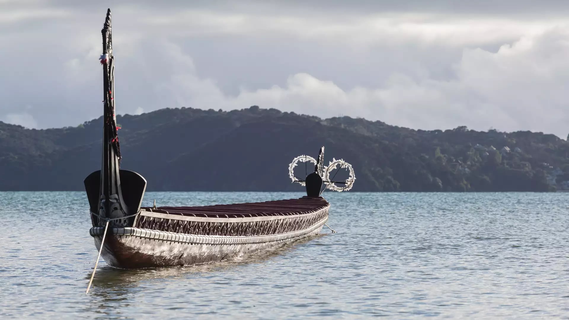 Traditional carved waka boat on calm water with hills and cloudy sky in the background Credit: Waitangi Treaty Grounds
