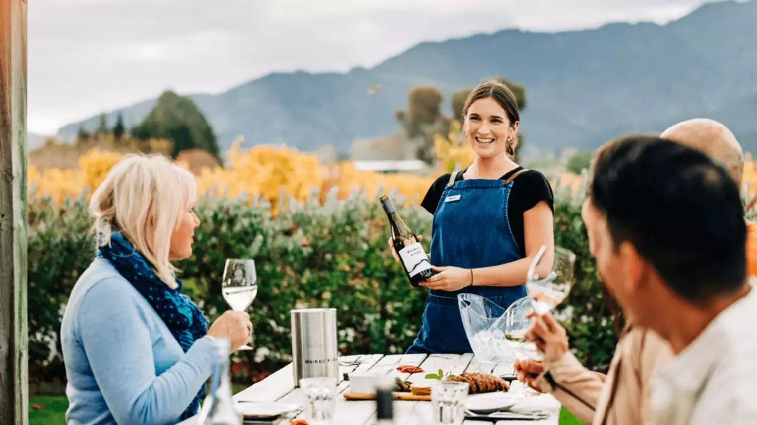 People enjoying outdoor wine tasting with a smiling host pouring wine at a vineyard with mountains behind.