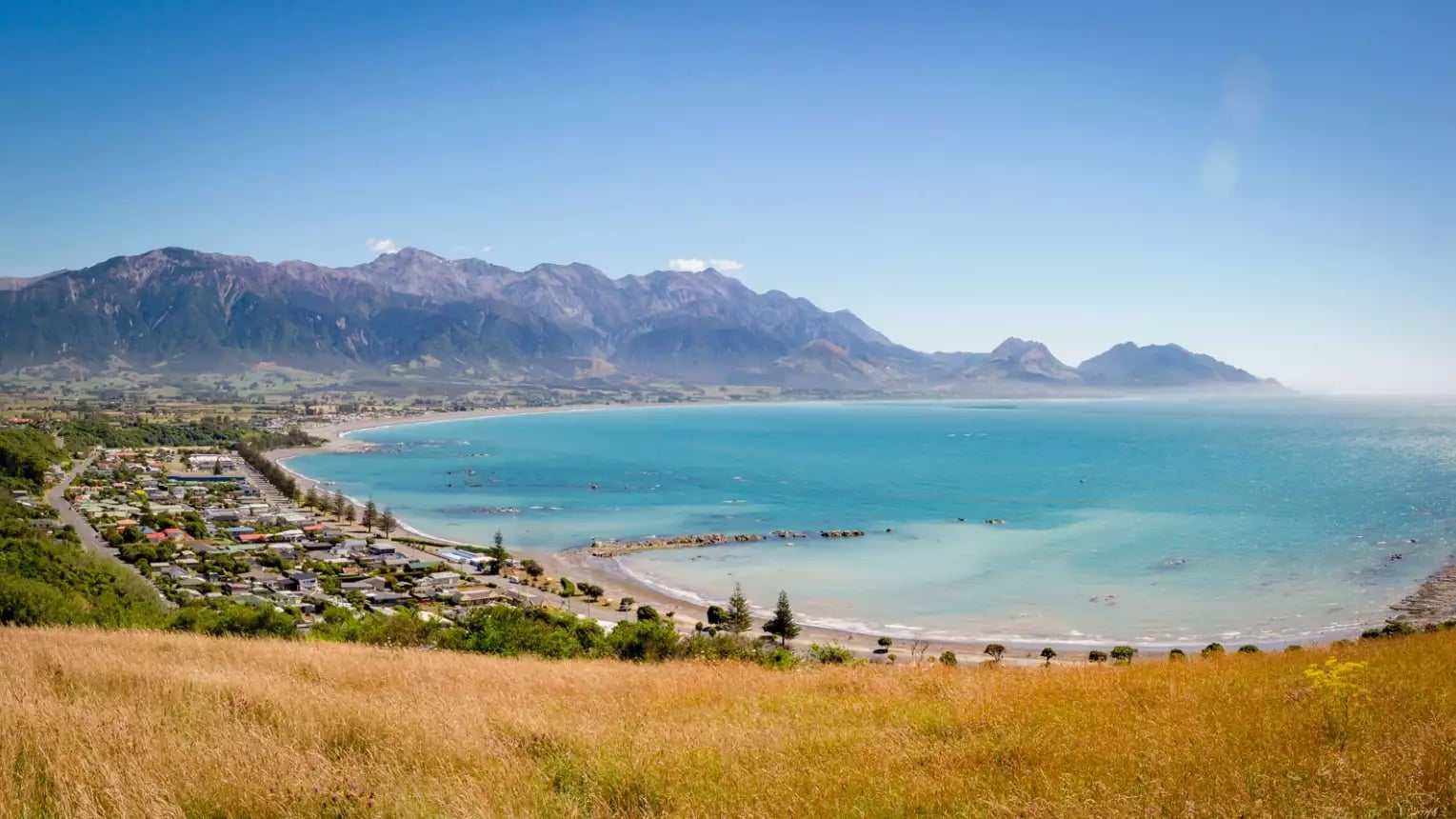 Coastal bay with clear blue water, town by the shore, mountains in background under a blue sky.