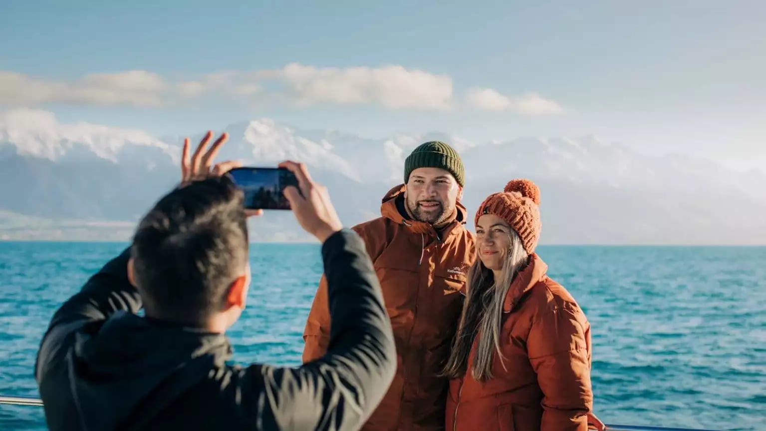Couple in warm jackets and beanies posing by a lake with snowy mountains behind.