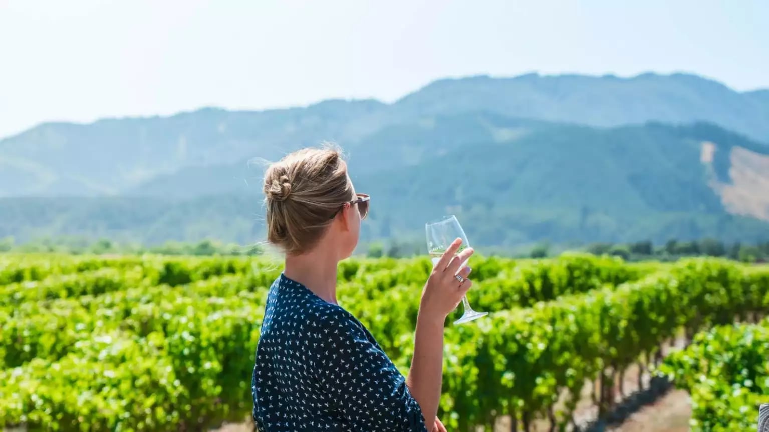 Woman holding a glass of white wine standing in a green vineyard with mountains in the background.
