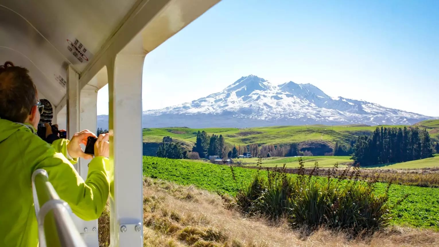 Tourists on a train taking photos of snow-covered Mount Ruapehu against blue sky and green fields.