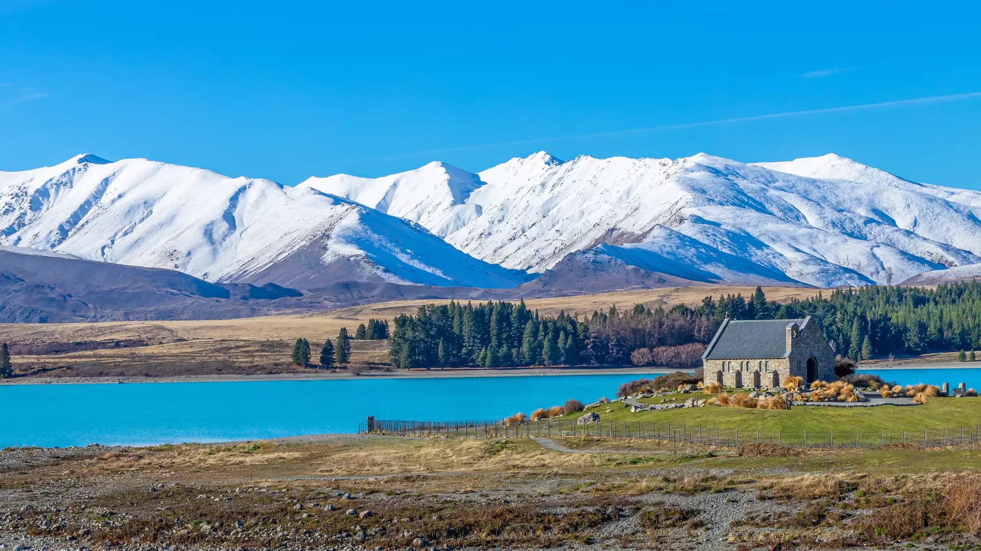 Stone church by bright blue lake with snow-capped mountains in background under clear sky.