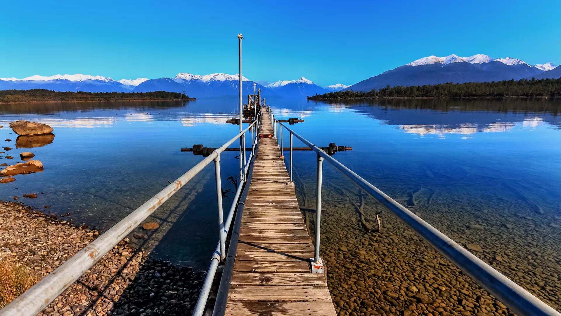Wooden jetty stretching into a lake with clear water and snowcapped mountains under a bright blue sky.
