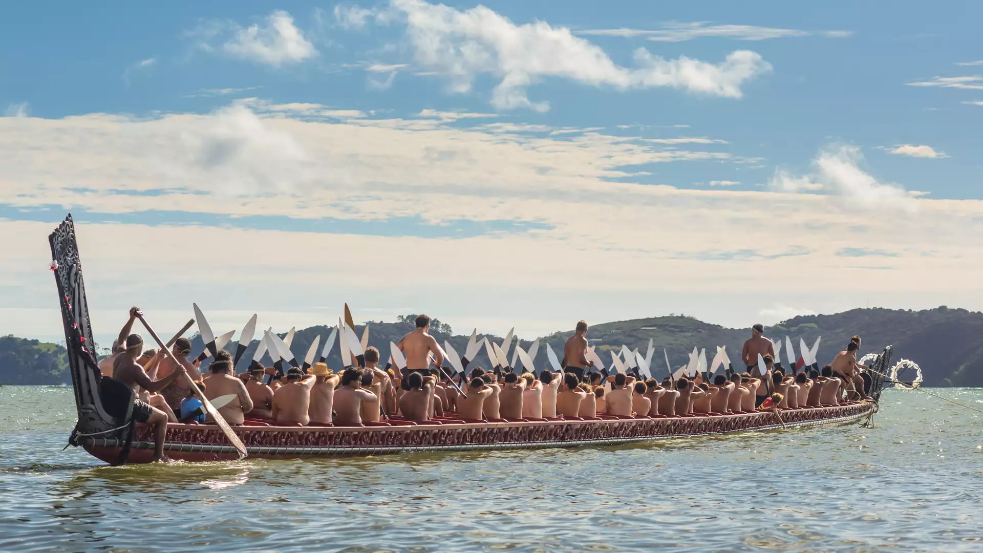 People paddling a traditional waka canoe on water with hills and blue sky in background.