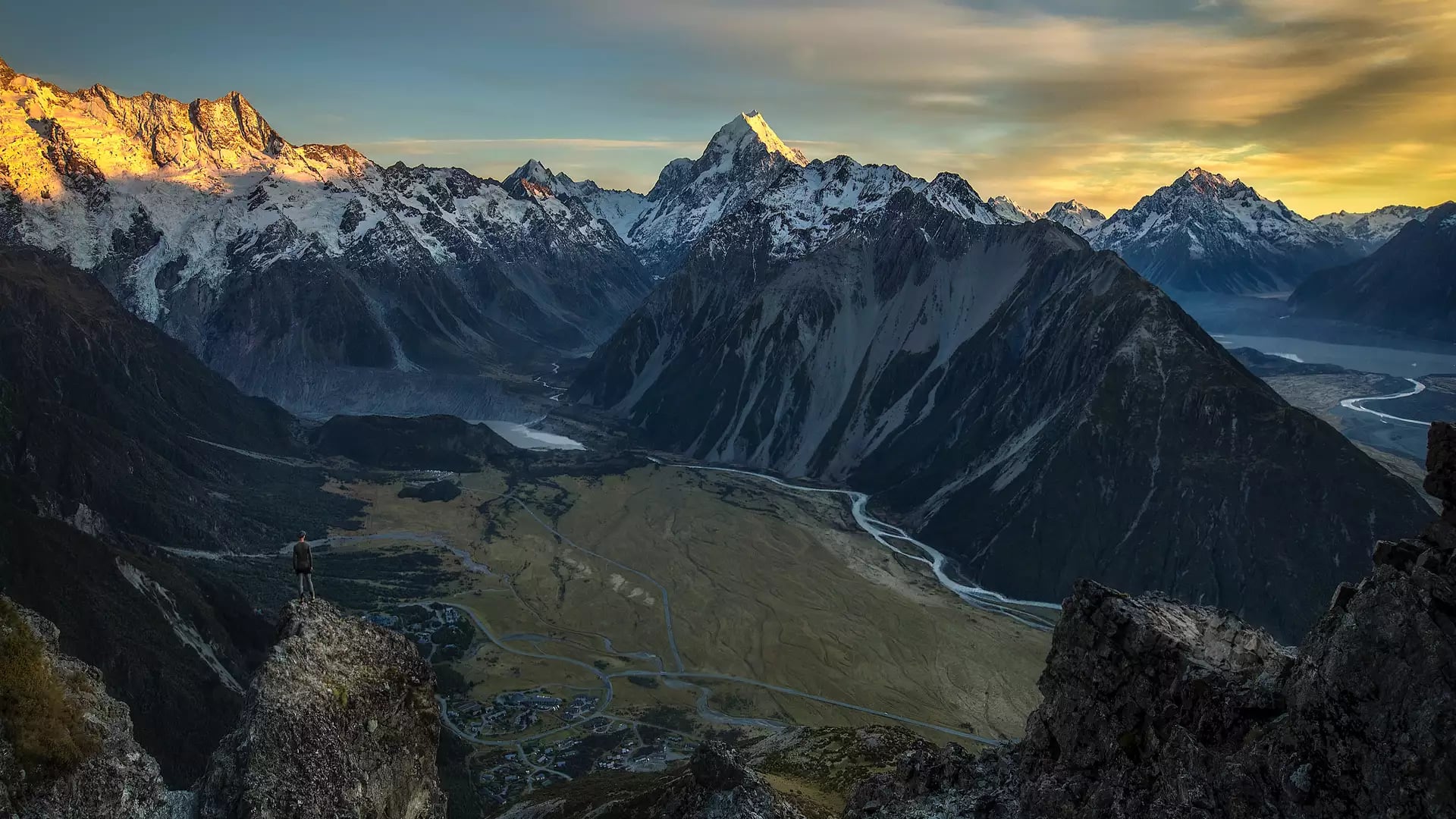 A high-altitude view of a person standing on a rocky outcrop overlooking a vast glacial valley, with the sun-drenched peak of Aoraki / Mount Cook in the distance under a golden sky. Credit: Mackenzie Tourism - Lee Cook