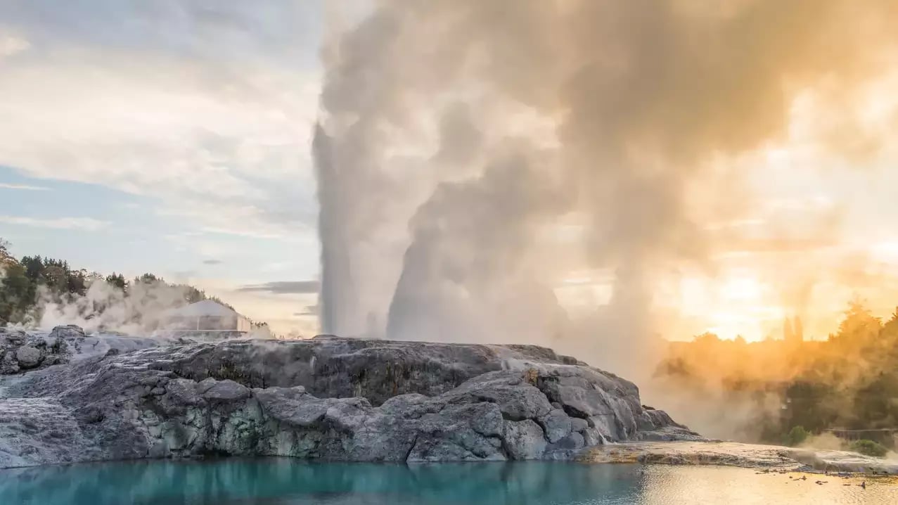 The Pōhutu Geyser erupts with a massive plume of steam and water behind a rocky silica terrace, reflected in a calm turquoise pool under a golden sky.