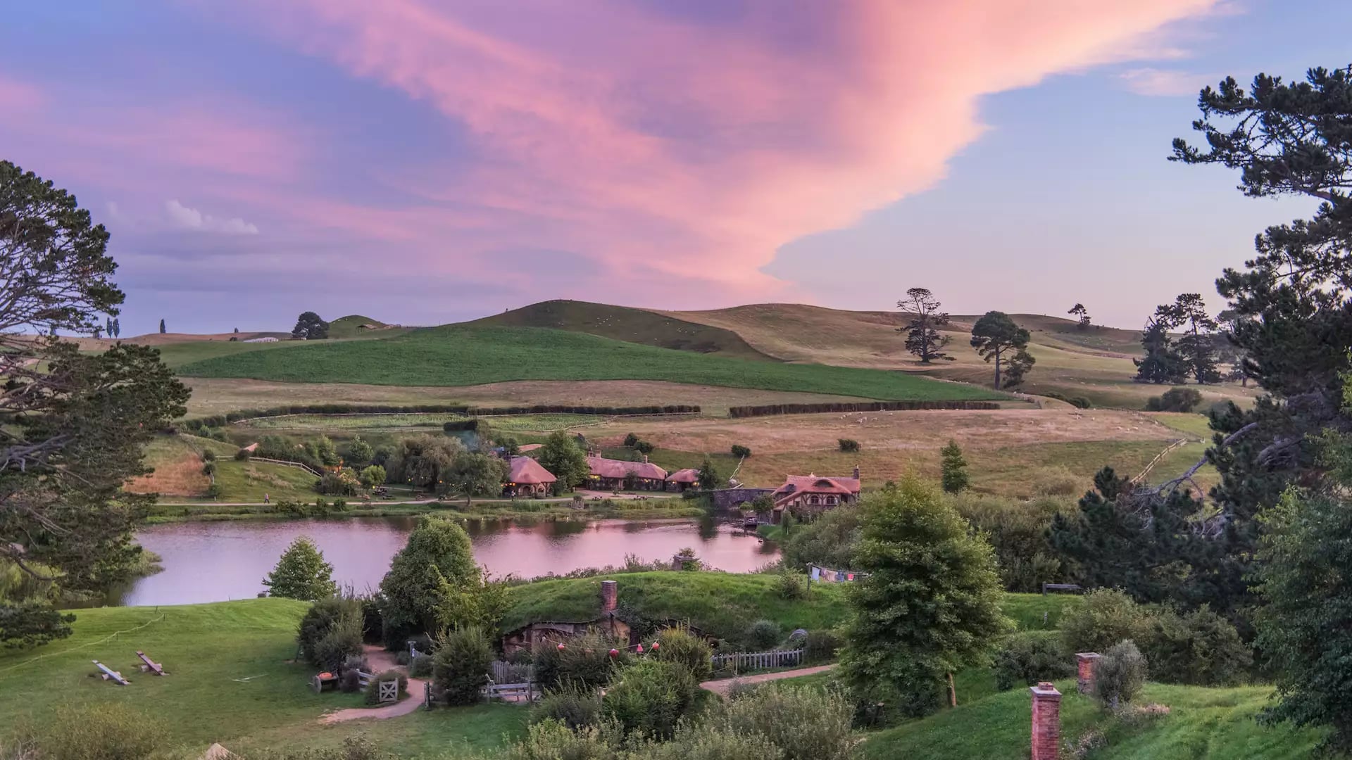 Scenic Hobbiton Movie Set with green hills, lake, trees, and buildings at sunset.