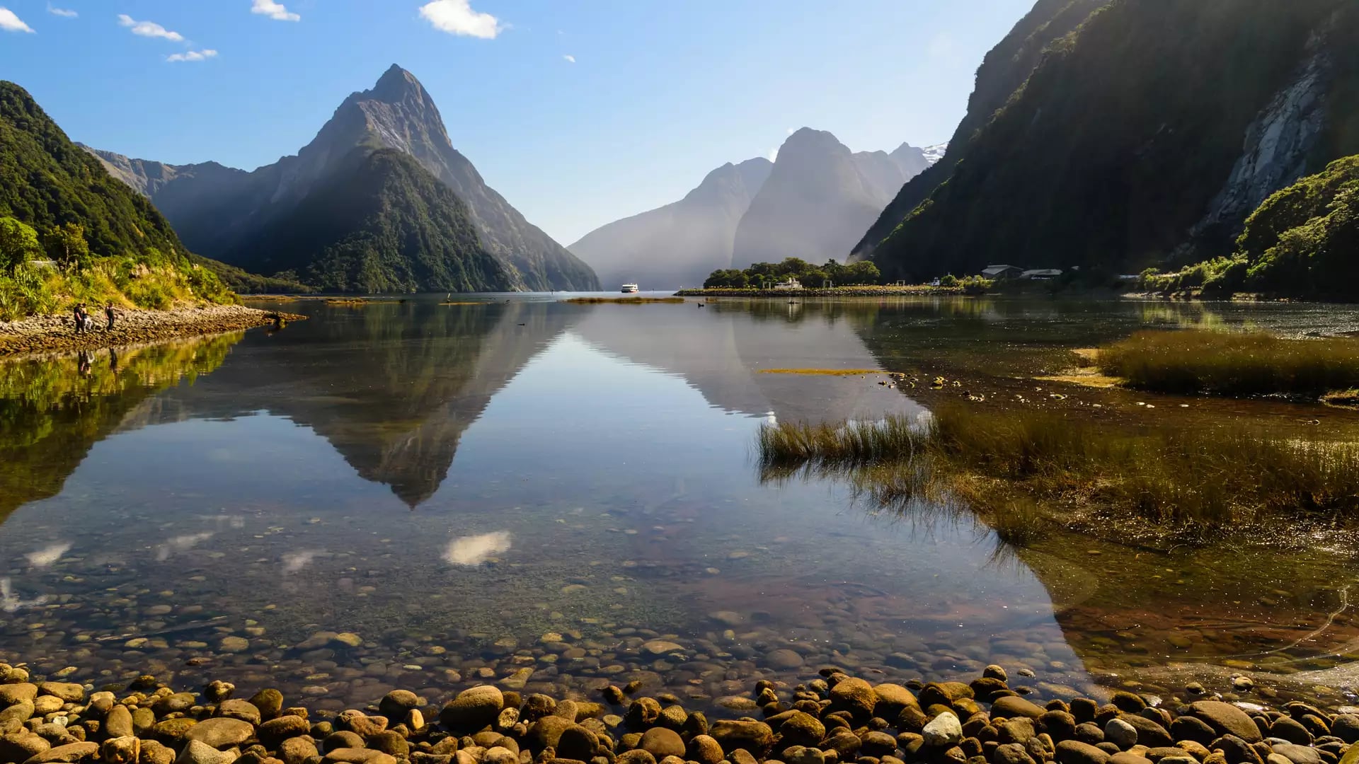 Milford Sound calm water reflecting mountainous landscape with rocky shore under blue sky.