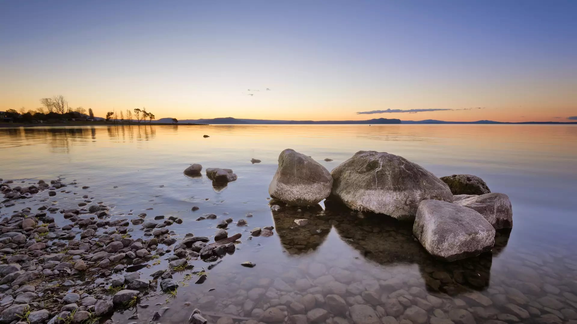 Sunset over Lake Taupo with rocks in the water and calm reflections.