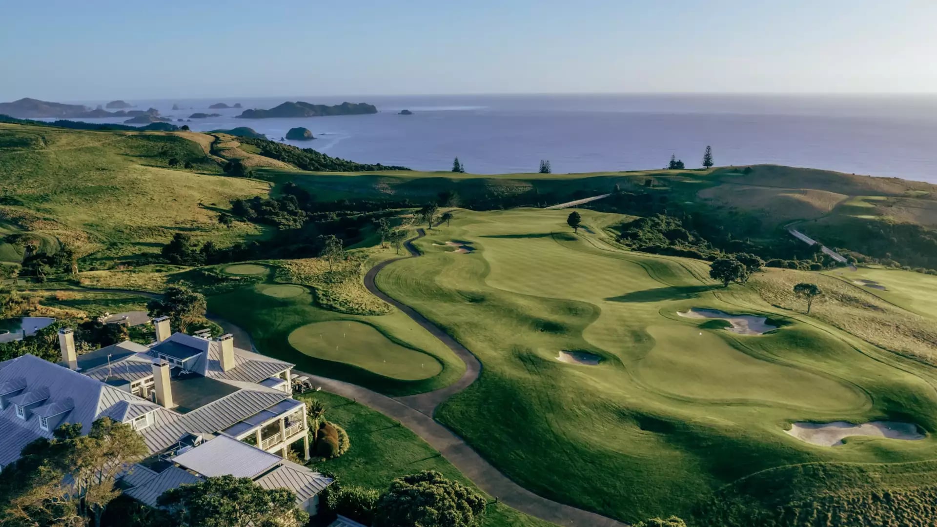 An aerial view of the lush green fairways and bunkers at Kauri Cliffs golf course, overlooking the Pacific Ocean and offshore islands.
