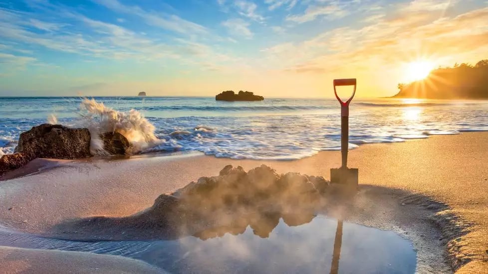 Shovel in sand on beach with sunrise, waves, and rocks nearby.
