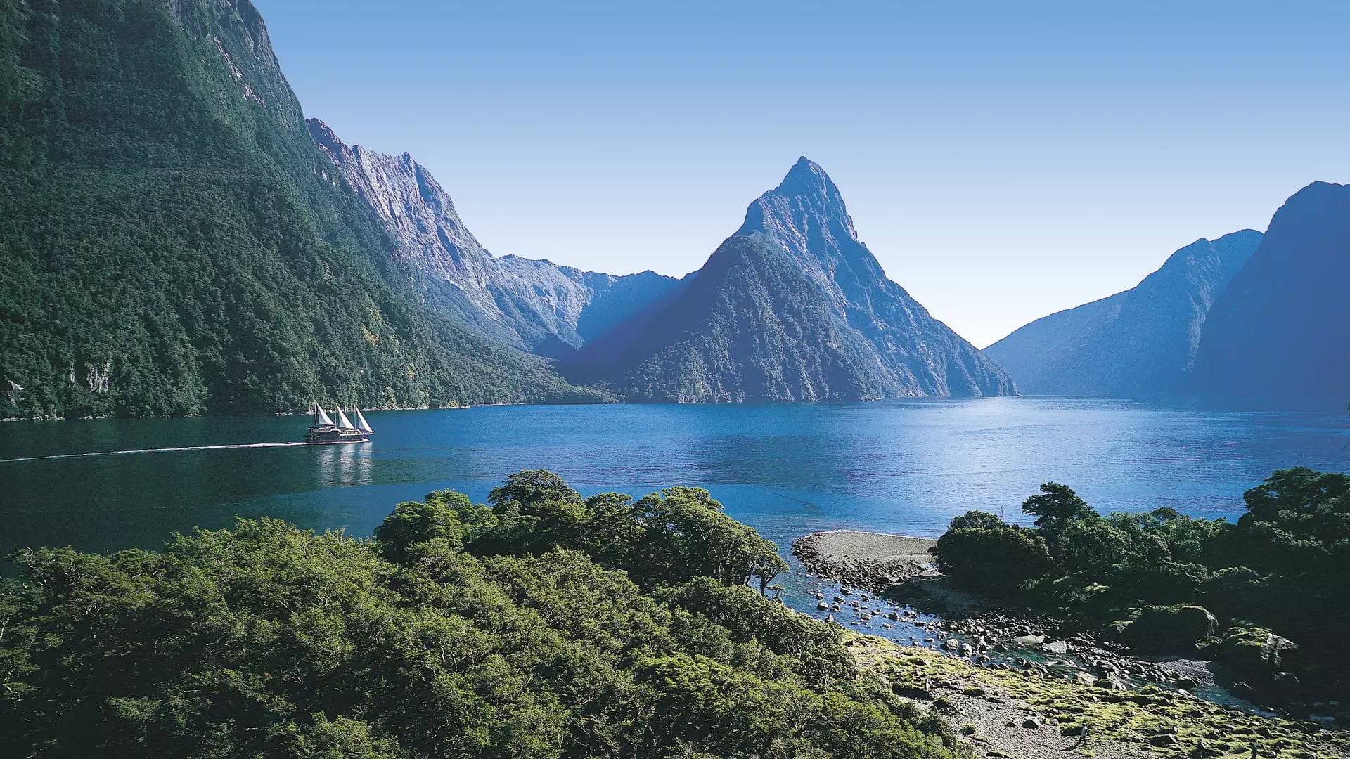 Sailboat on calm blue water surrounded by towering green mountains under clear sky.