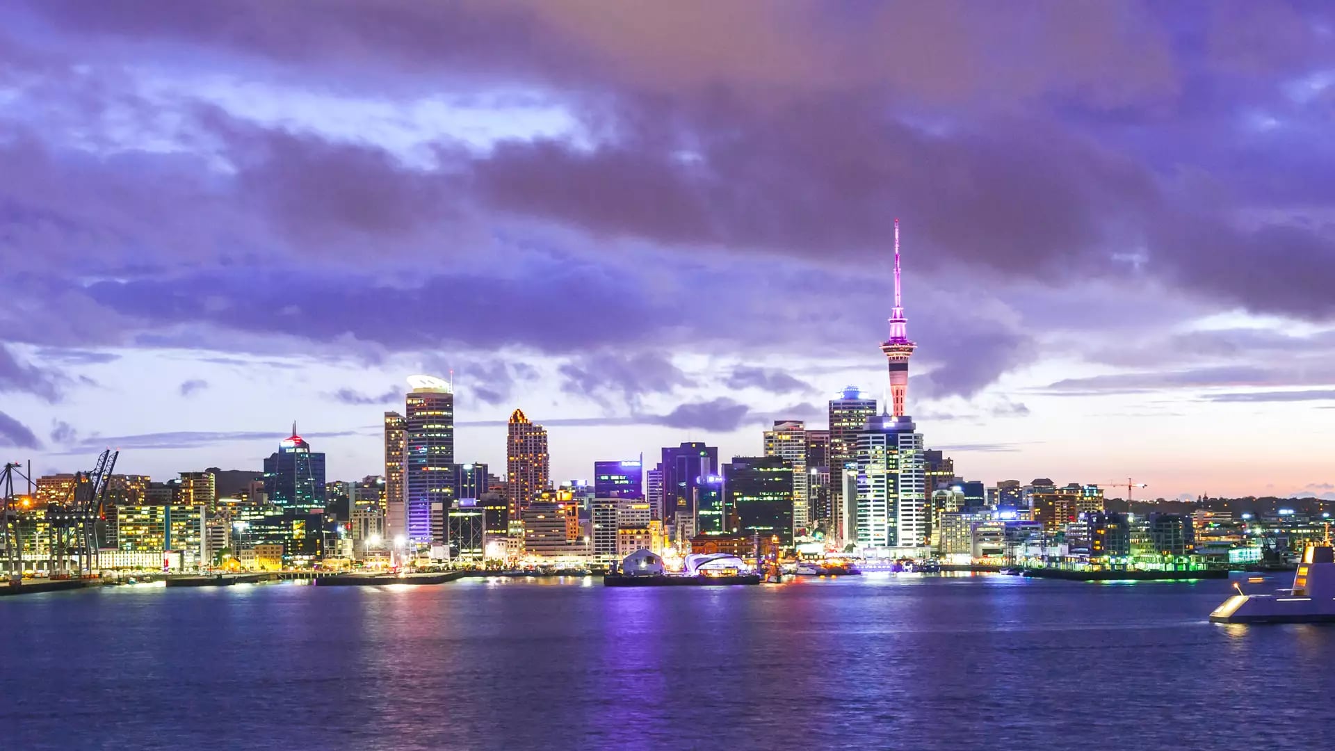 Auckland city skyline at twilight with purple clouds and city lights reflecting on the harbour water.