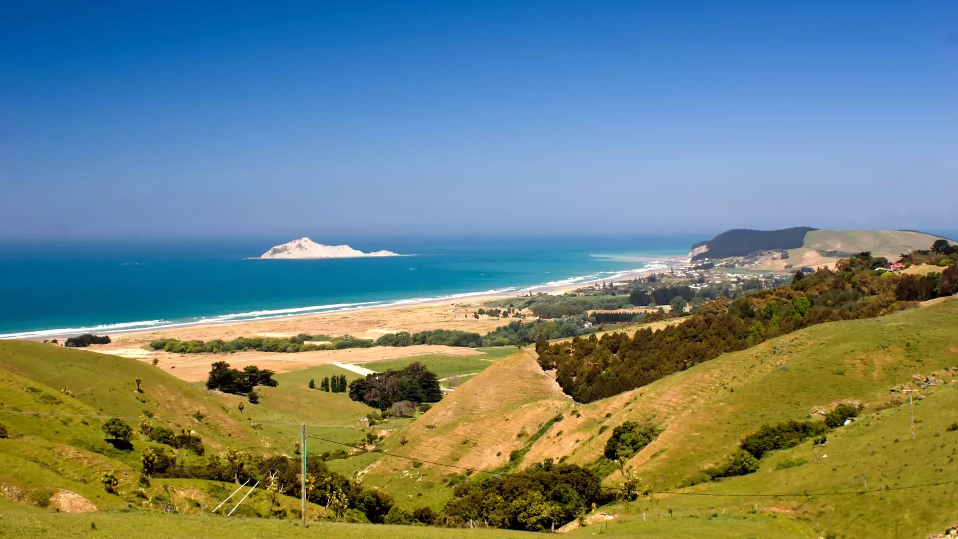 View of green hills, sandy beach, ocean, and a small island off the coast under a clear blue sky.