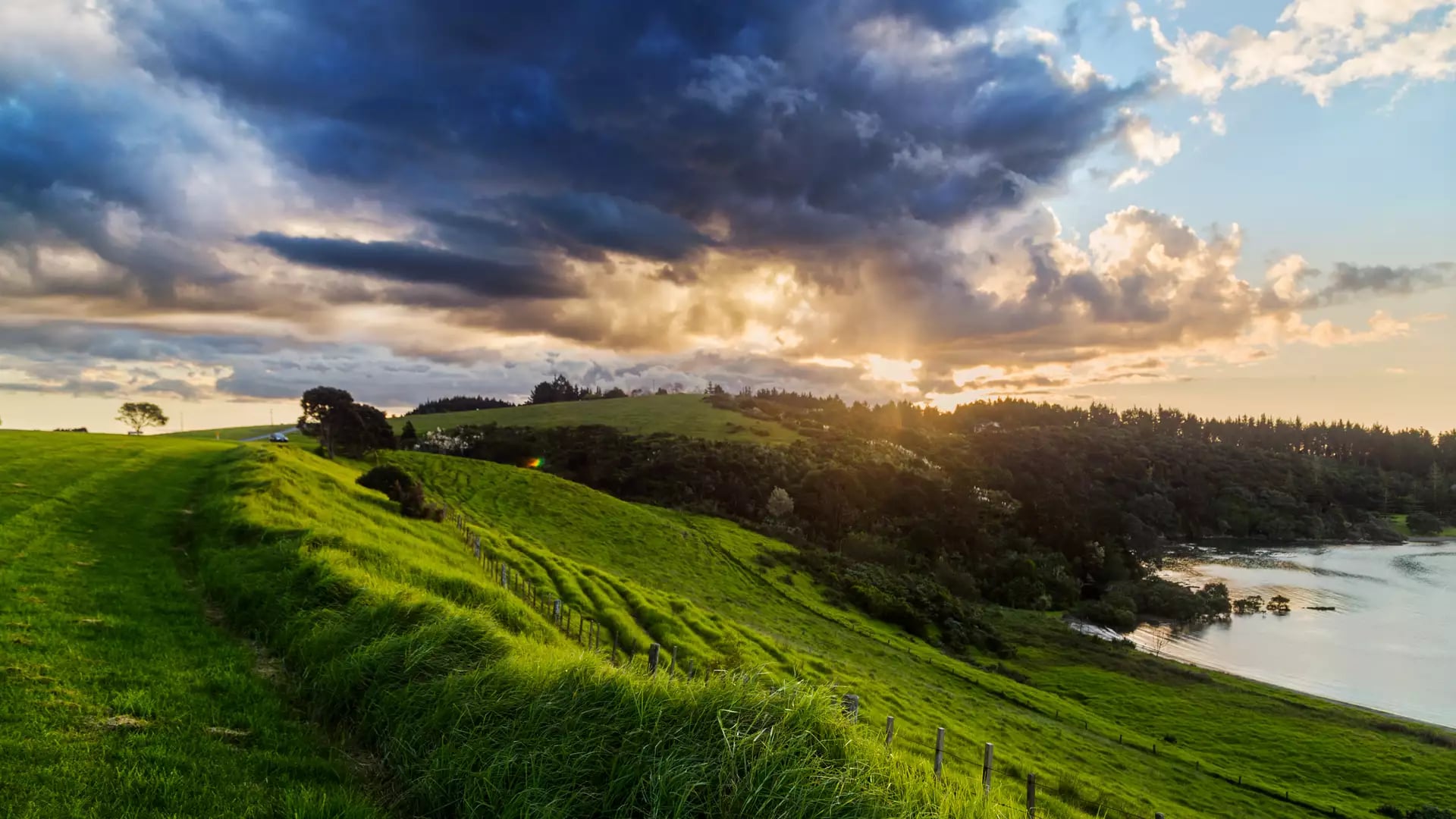 Lush green hills with a wooden fence overlooking a calm bay under a sky with dark clouds and sunbeams.