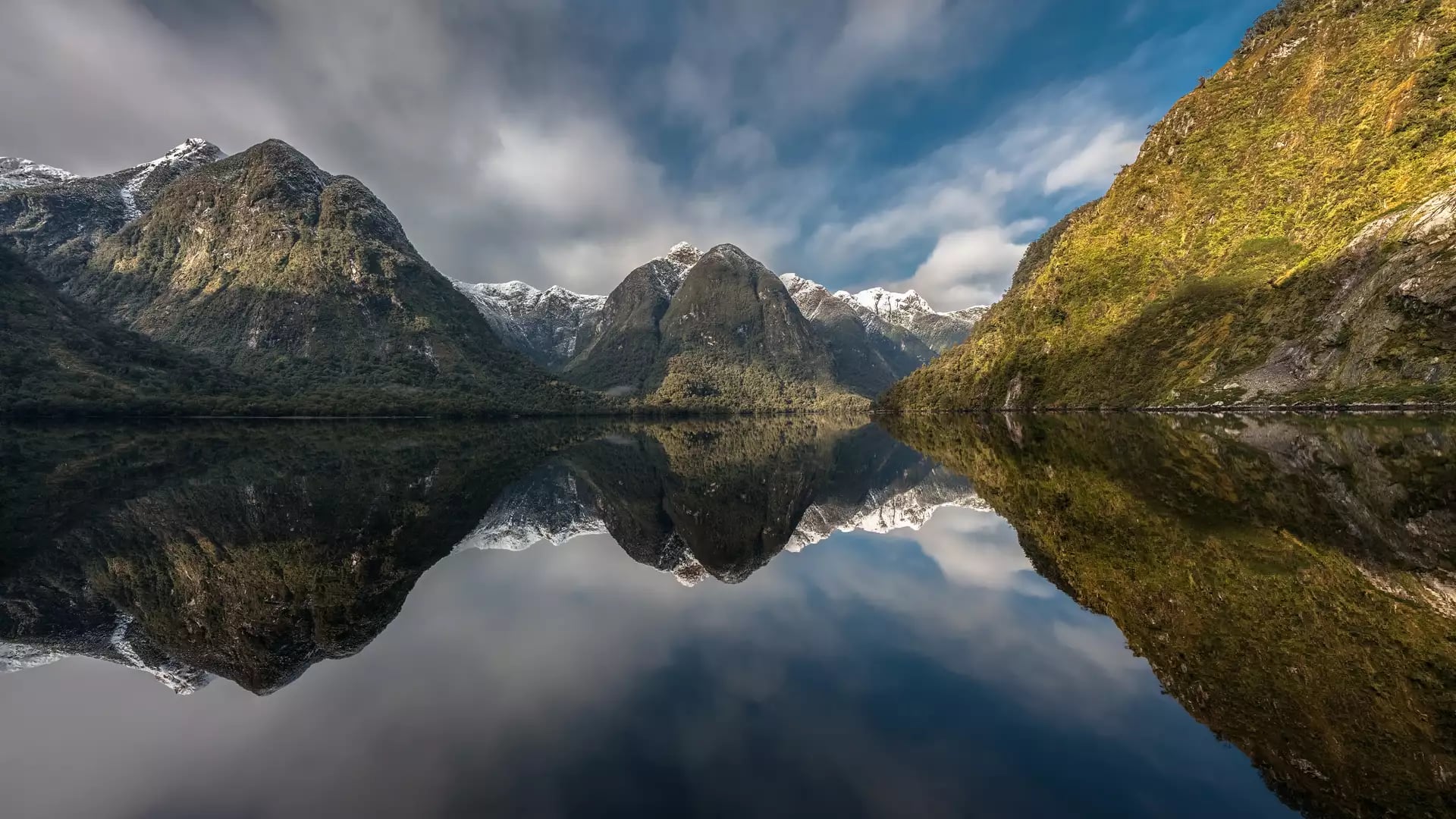 Mountain peaks reflecting on calm lake water with partly cloudy sky above.