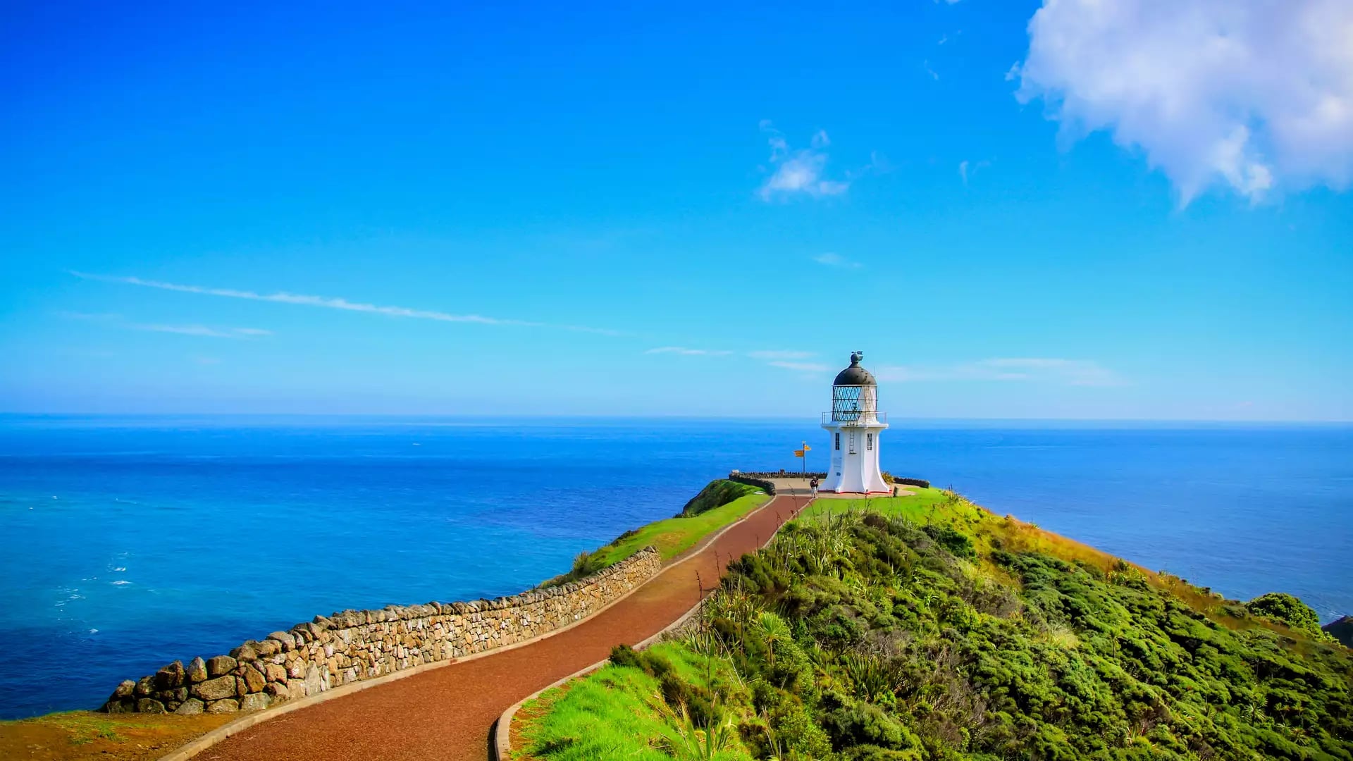 White lighthouse atop a green cliff with a pathway leading to it under a bright blue sky.