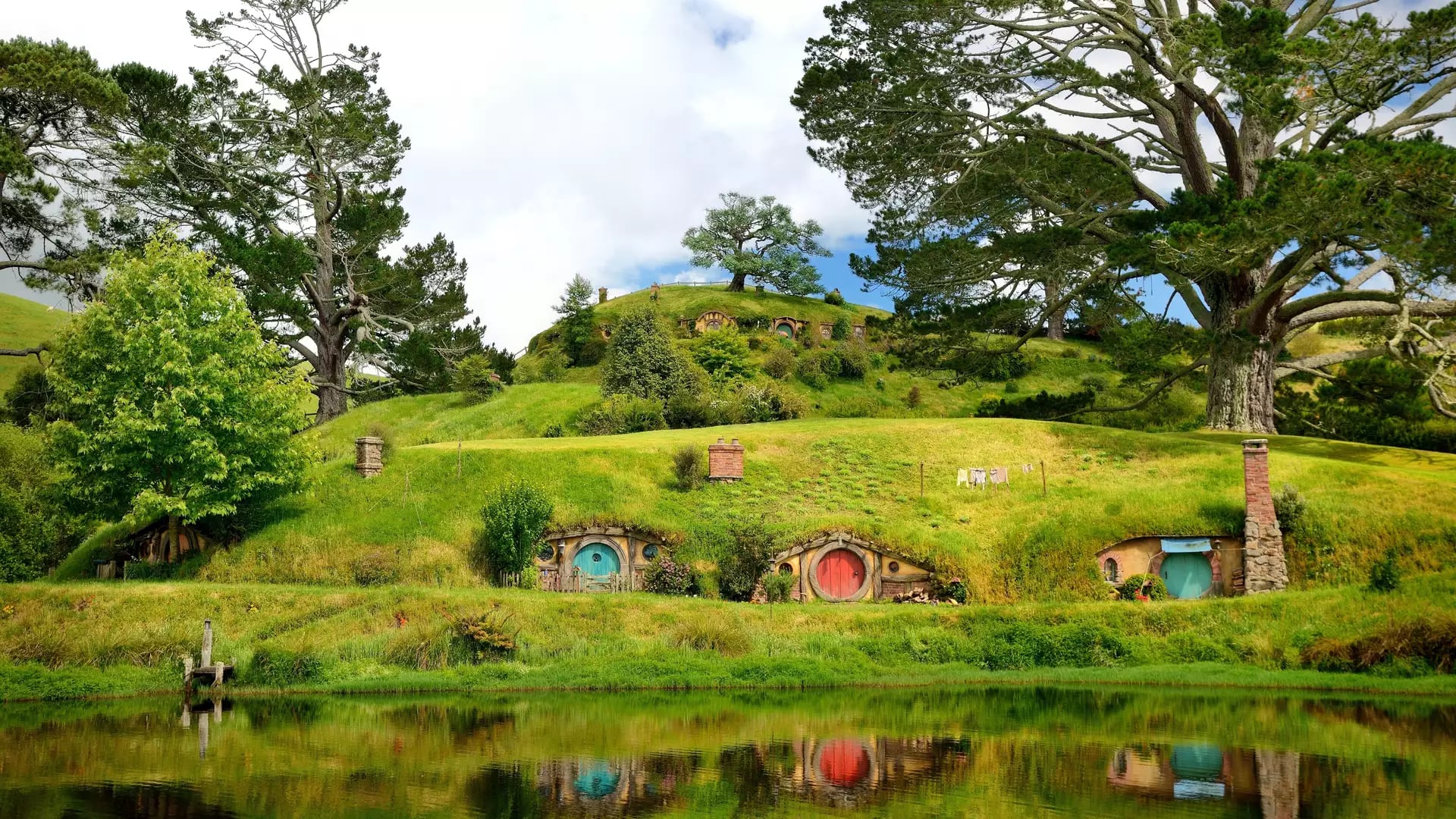 Colorful hobbit house doors built into green hills with large trees and a pond reflecting the scene
