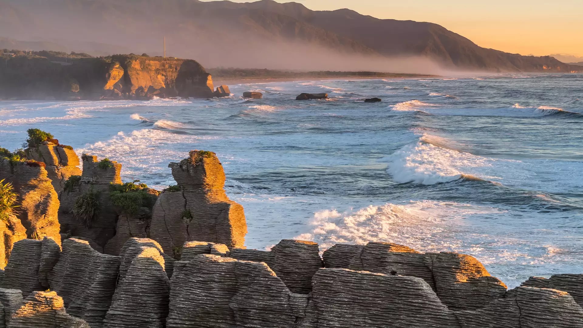 Pancake Rocks coastal rock formations at sunset with waves and mountains in the background