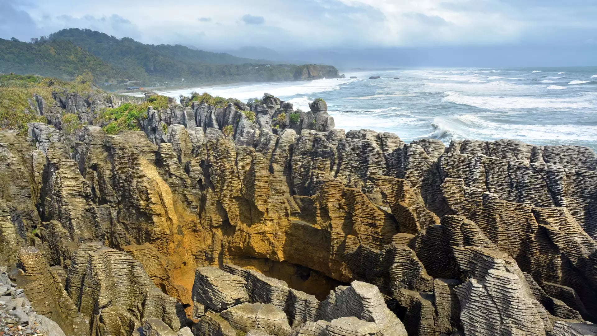 Layered limestone rock formations known as Pancake Rocks with ocean waves and green hills in the background