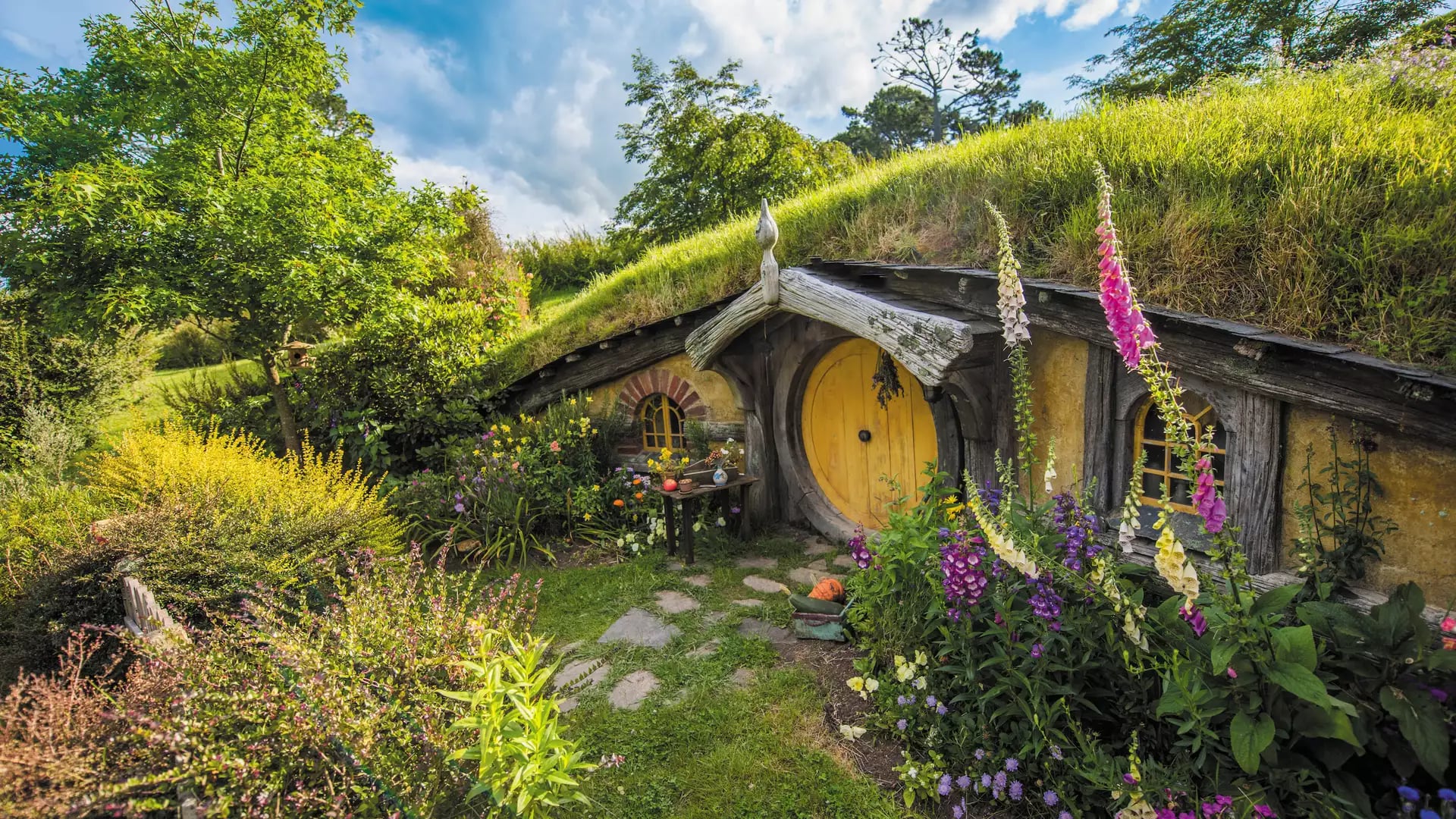 Hobbiton hobbit house entrance with round yellow door surrounded by colorful flowers and greenery under a blue sky.