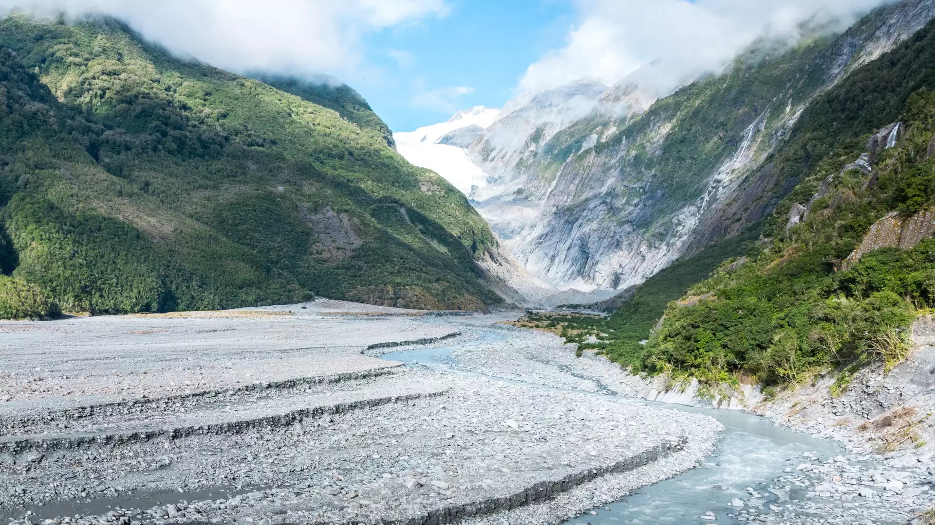 Franz Josef Glacier flowing through mountain valley with river and green forest under blue sky