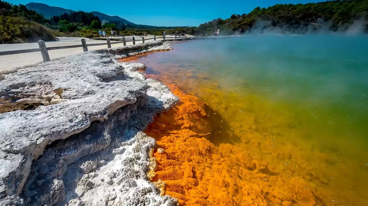 A close-up view of the vibrant orange mineral edge and turquoise steaming water of the Champagne Pool, with a wooden boardwalk and visitors in the background.