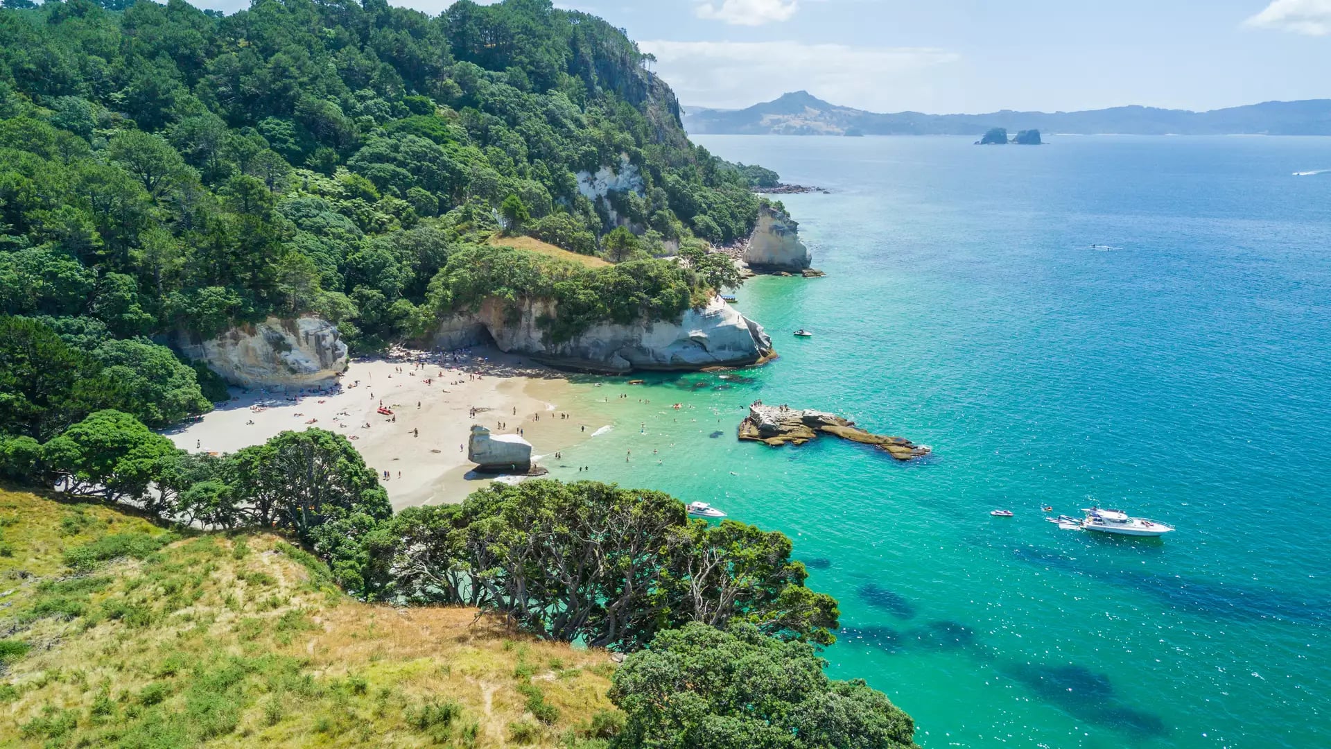 Aerial view of a sandy beach with turquoise water and boats near forested cliffs at Cathedral Cove.