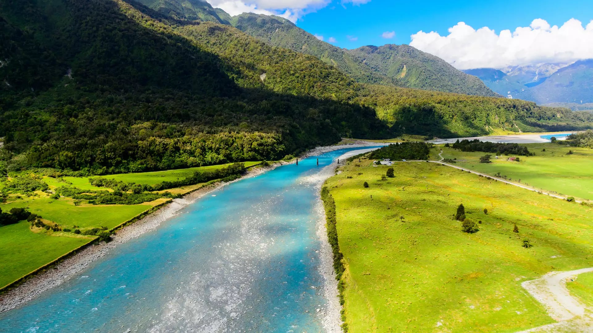 Blue river flowing through green valley with forested mountains in background under a partly cloudy sky.
